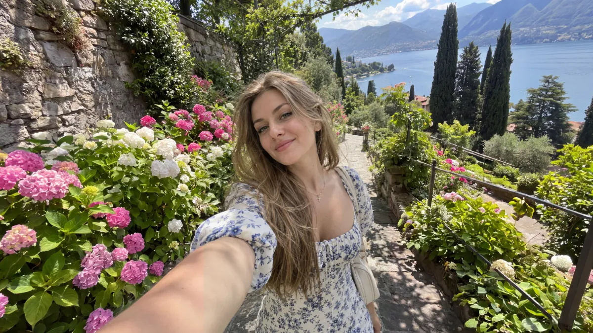 The same selfie widened to landscape: hydrangeas and a stone wall to the left, cypresses and the lake to the right